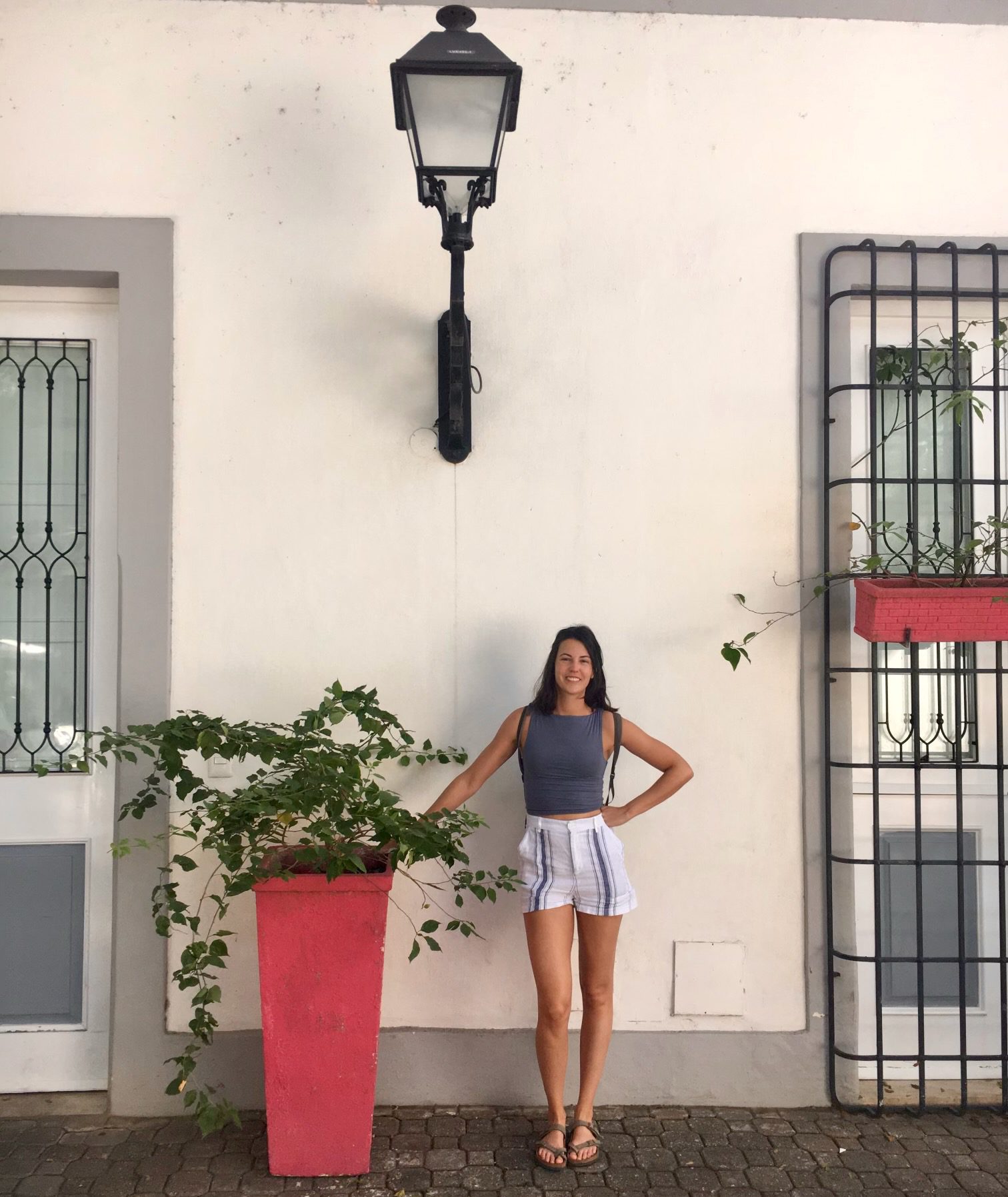 Una mujer en pantalones cortos y camiseta de tirantes posa ante una pared blanca junto a una alta jardinera