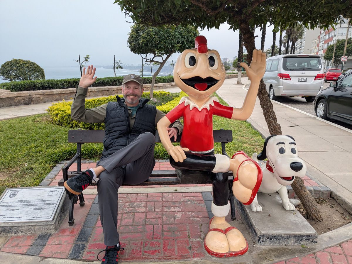 Scott sits on a park bench with a statue of Condorito... both wave at the camera