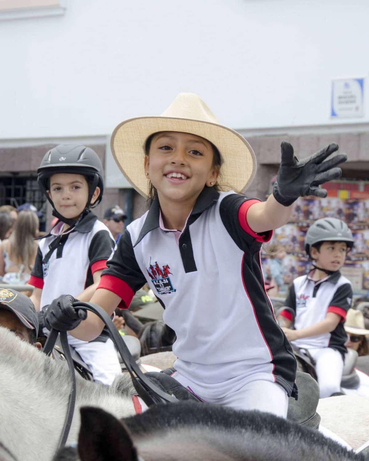 A young girl in riding gear and black gloves reaches toward the crowd with a smile on her face