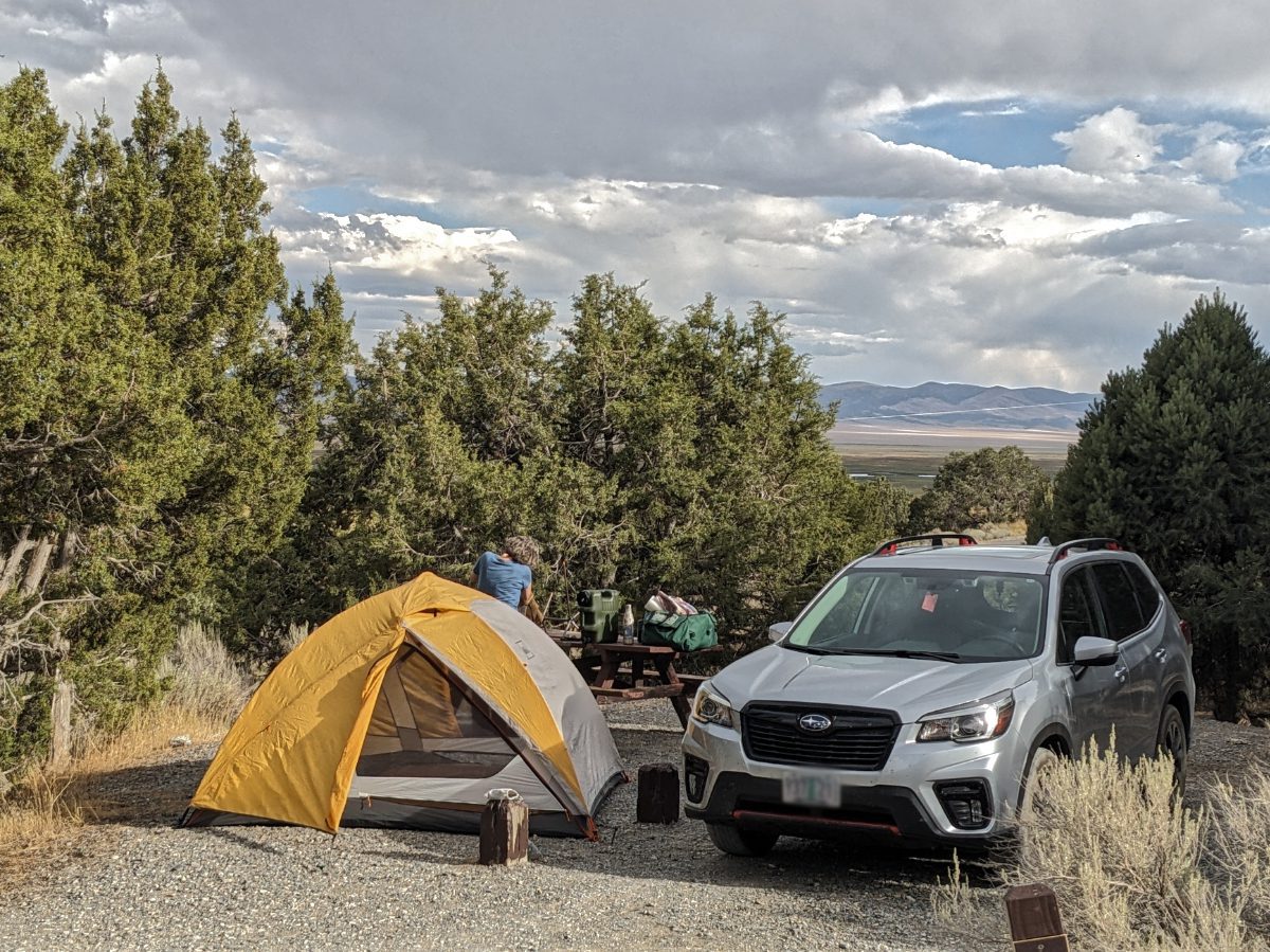Car camping on a gravel pad, view of tent and gray Subaru, woman working in camp kitchen in background, all with a gorgeous view of pine forest, wetlands, and distant hills.