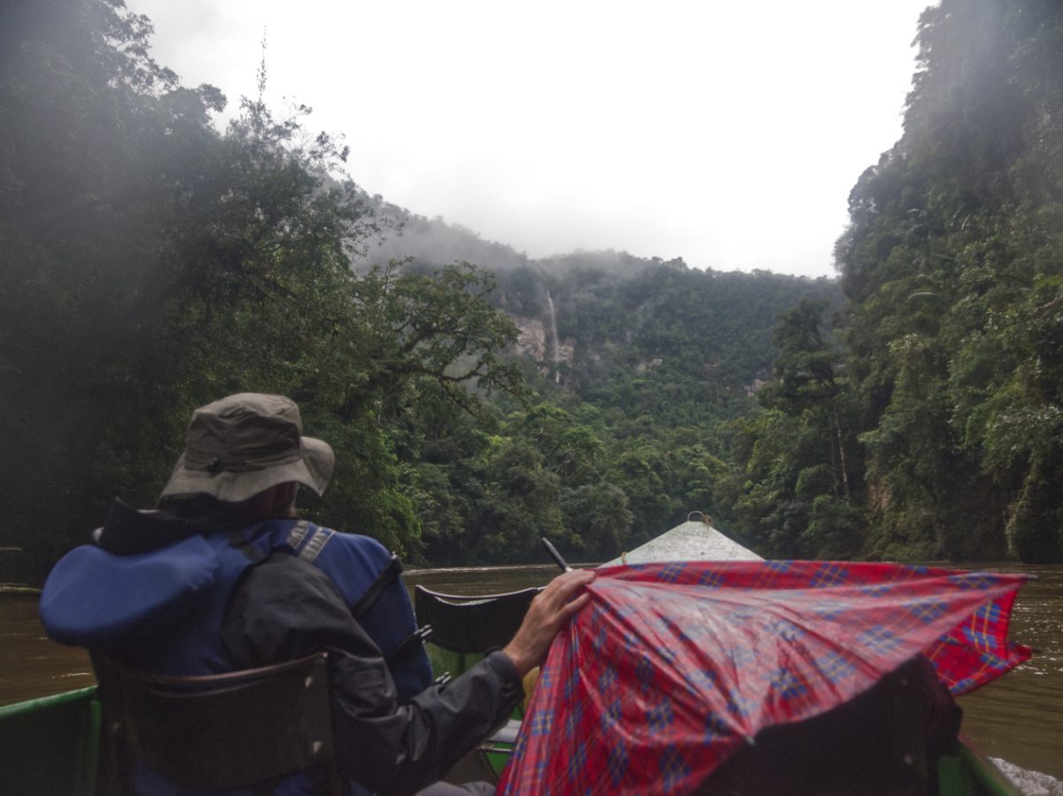 A man on a boat on a river bordered by rainforest and a waterfall places his hand on an umbrella protecting a backpack.
