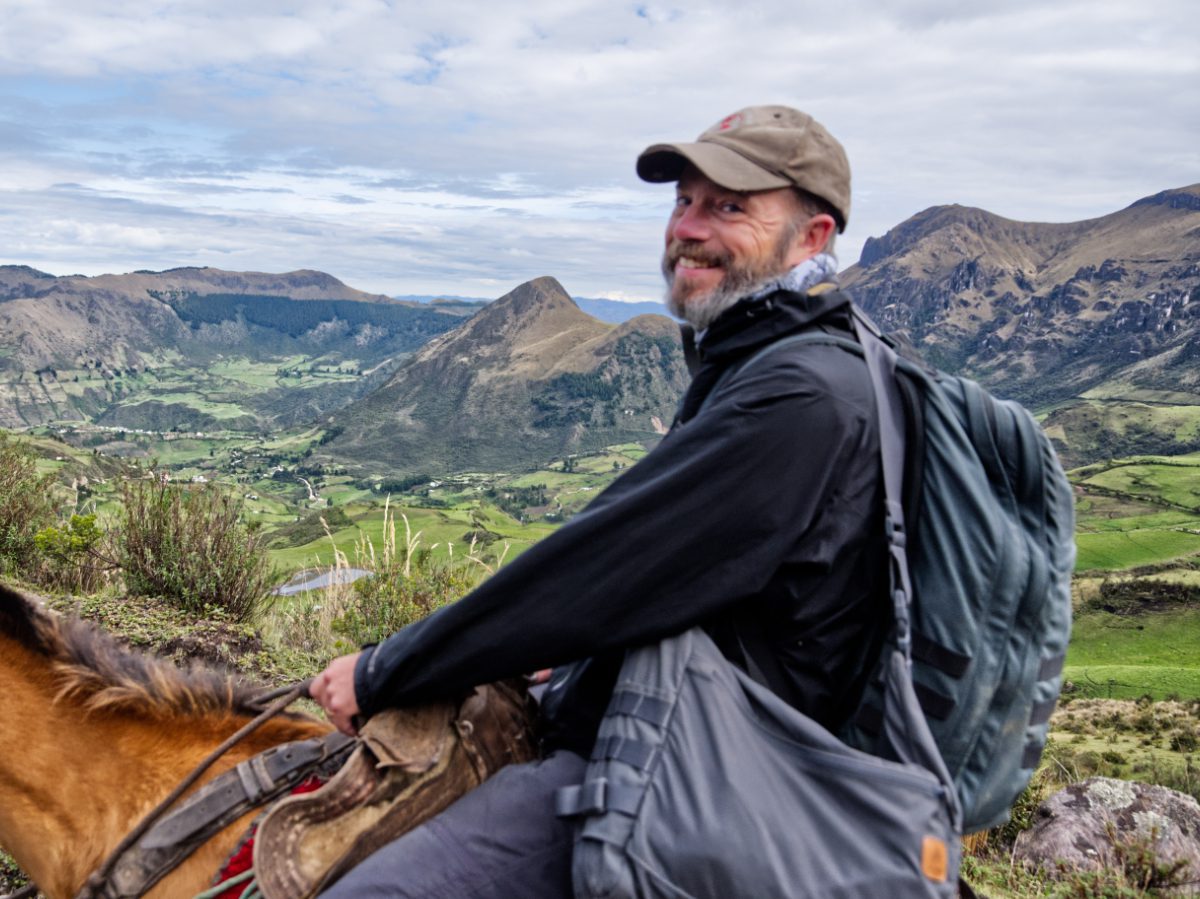 A man wearing a backpack while mounted on a horse turns and smiles at the camera