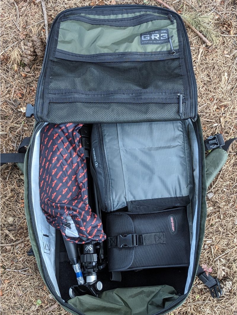 A GR3 backpack zipped open to show tripod, binoculars, and camera gear stowed inside.
