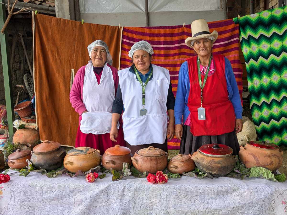 Three women dressed in aprons smile at the camera while standing behind a long table full of clay pots.