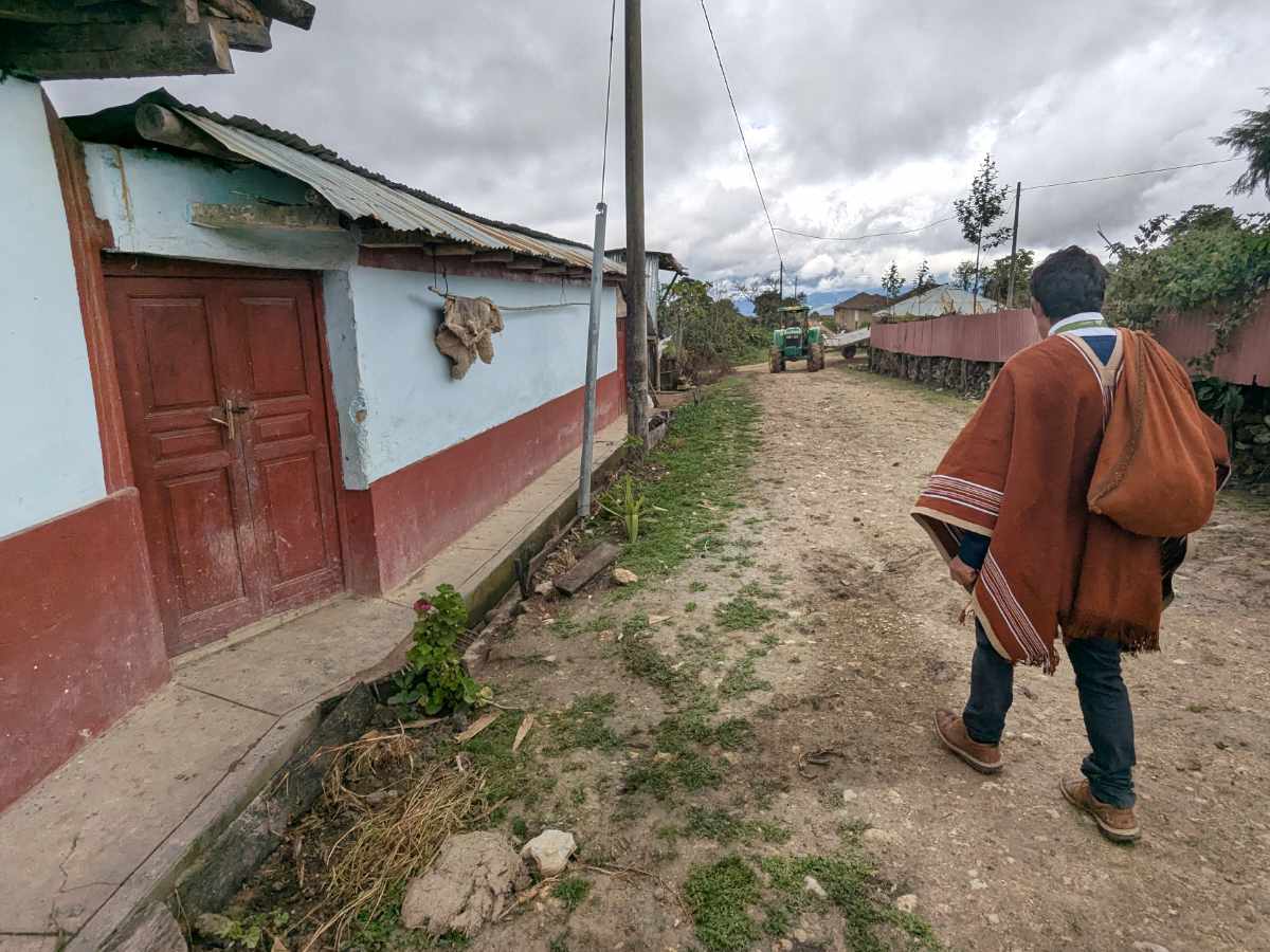 A man in a deep rust colored poncho walks on a dirt road by a house painted light blue with oxblood red trim.